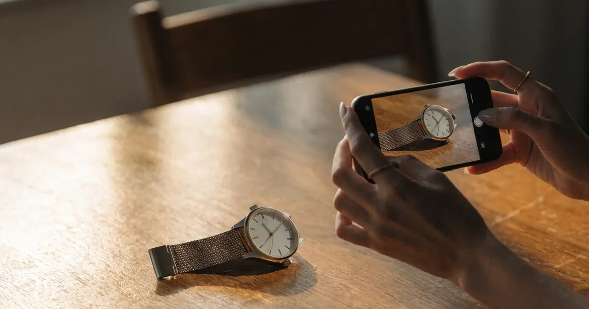 Une main photographie avec un téléphone une montre-bracelet posée sur une table en bois ; la montre apparaît aussi à l'écran du téléphone.
